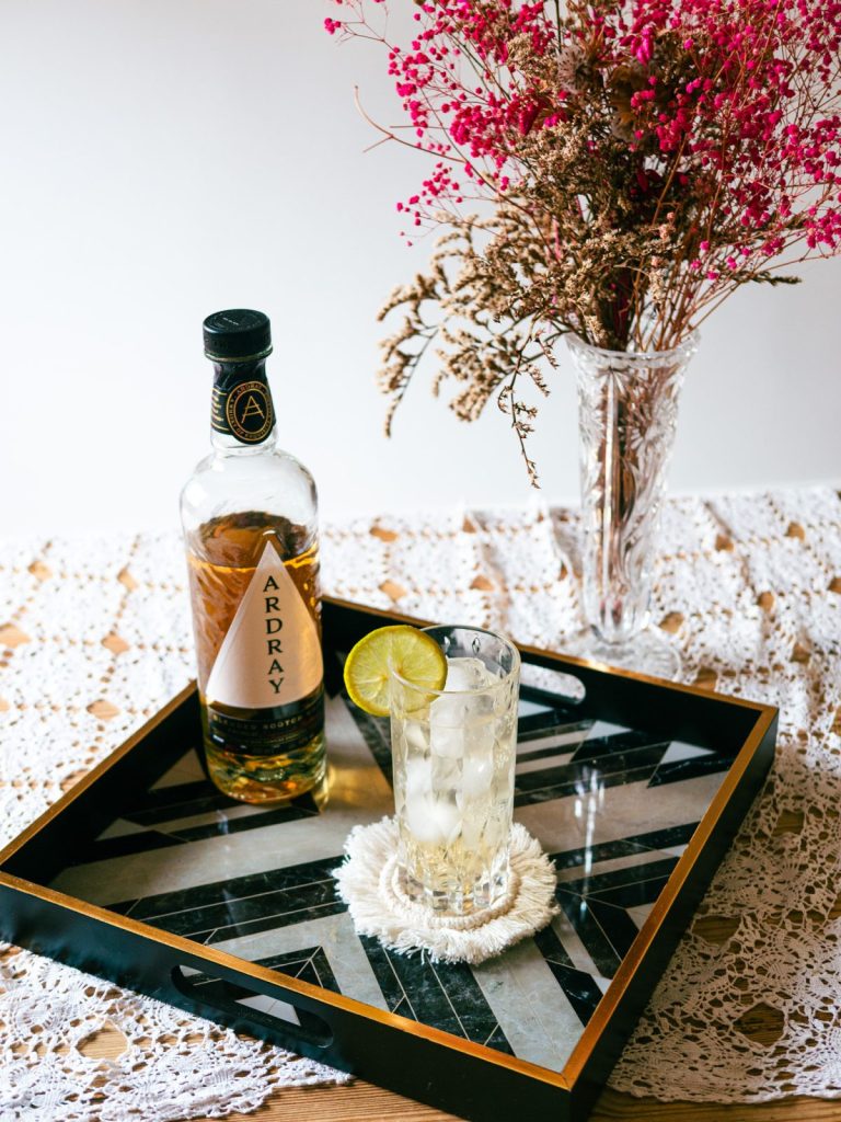 A tray with a highball and whisky bottle with dried flowers in the background and a lace table cloth 