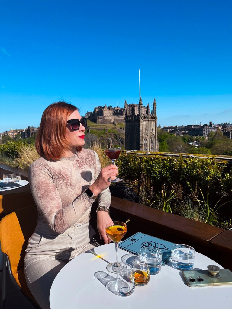 A woman holding a cocktail on a roof top terrace over looking the Edinburgh Castle.